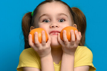 A small and smiling girl is holding two tangerines on a blue background.