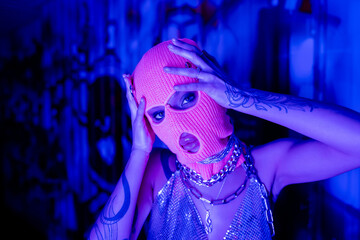 passionate tattooed woman in silver neck chains looking at camera while touching knitted balaclava in blue and purple light.