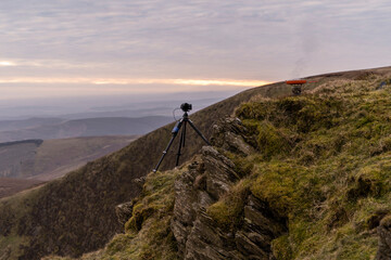 A camera and tripod on a mountain cliff edge with frying pan camping setup