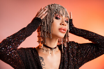 portrait of tattooed woman in black lurex dress touching wig with shiny rhinestones on coral pink background.
