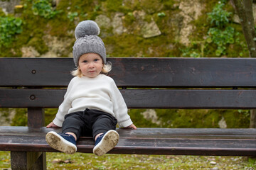 Cute joyful blonde kid sitting on a bench outside in a park after playing in dirt. Happy child wearing a hat outside on a spring day. Little boy resting on a bench with his dirty shoes.