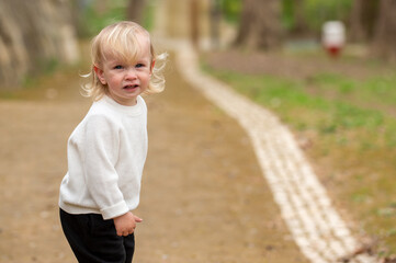 Fototapeta premium Cute blonde kid in a white sweater is walking around a park. Interested caucasian baby boy alone on a sidewalk on a chilly day. A boy in a white sweater exploring the world outdoors.