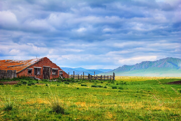 Obraz premium Beautiful scenery with old abandoned farm. Ruined building with a wooden fence and green field at the background of mountain range and cloudy sky in Khakassia, Southerh Siberia, Russia