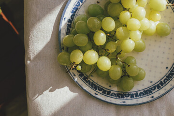 Fresh grapes in sunlight on ceramic plate flat lay. Healthy food aesthetics. Summer fruits in light on soft linen background. Summertime in countryside, moody banner