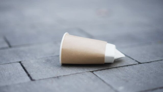 Close-up paper coffee cup on pavement with male feet passing leaving. Unrecognizable careless man stepping on trash walking away on city street. Contamination concept