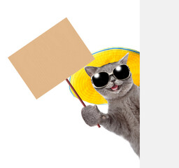 Happy cat wearing  sunglasses and summer hat looks from behind empty board and shows empty placard. isolated on white background