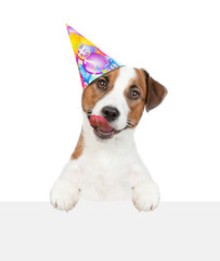 Licking lips Jack russell terrier puppy wearing party cap looks above empty white banner. isolated on white background