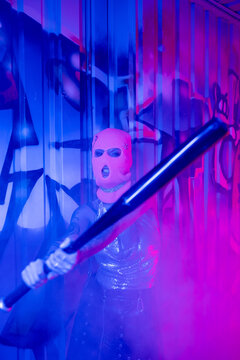 aggressive woman in balaclava grimacing while holding baseball bat near graffiti in blue and purple light with smoke.