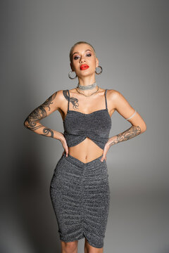 extravagant tattooed woman with short hair and bright makeup looking at camera while posing with hands on waist on grey background.