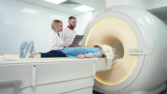 Woman doctor radiologist explains good results of CT scanning for young female patient, showing the snapshot with images, observing and analyzing CT scan in modern clinic beside modern CT scanner