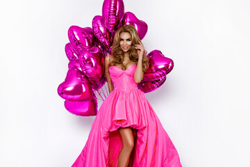 Beautiful woman in amazing long dress is holding pink balloons and is posing on white background. Elegant female model in gown dress. Elegance. Glamour.