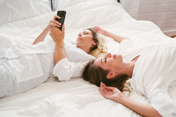 Obraz na płótnie Canvas Happy two friends Caucasian young women in white bathrobes. One looking at the camera and the other one using a smart phone while laying on the bed. LGBTQ couples leisure spa and treatment activities.