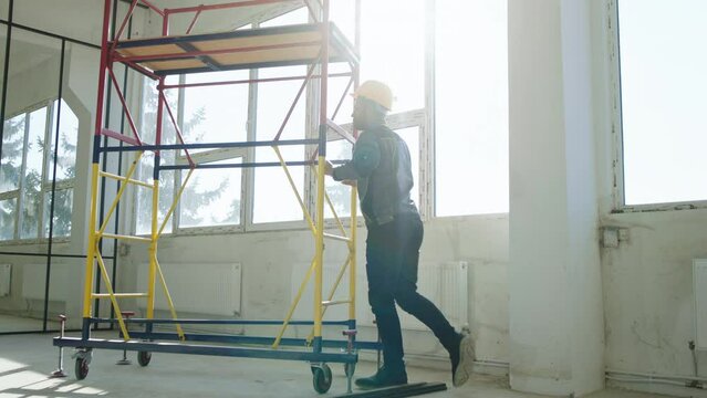 Good looking young multiracial constructor worker take the mobile ladder and rolling down through the construction site he wearing protective equipment and uniform