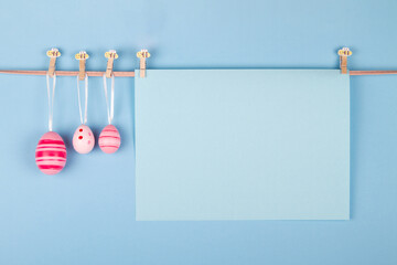 Blank pastel blue sheet of paper and three pink Easter eggs hang at rope on light blue background. Easter card with a designated area for writing