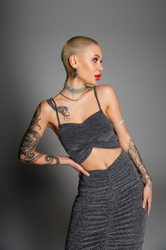 seductive woman in lurex crop top and necklaces looking away while posing with hand on hip on grey background.