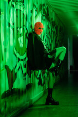 full length of sexy woman in stylish leather boots and orange balaclava standing near wall with graffiti in green lighting.