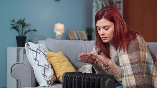 young woman wrapped in warm blanket suffers from cold in an unheated room and tries to warm her hands near electric heater