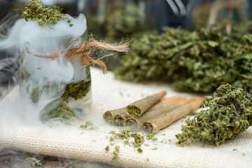 Marijuana joint pre-rolled cone paper with cannabis buds in a clear glass jar sit on a smoky floor