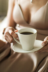 Beautiful young woman in a silk dress holds a cup of coffee. Close up view of hands with tea couple. Selective focus. Hotel and restaurant concept