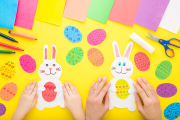 Mother and child hands with white smiling bunny and colorful eggs from paper on yellow table...