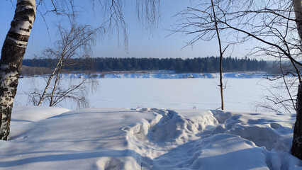Winter landscape with trees on a cliff and a view from a height of a frozen river or a field with snow on a cold sunny day with a blue sky
