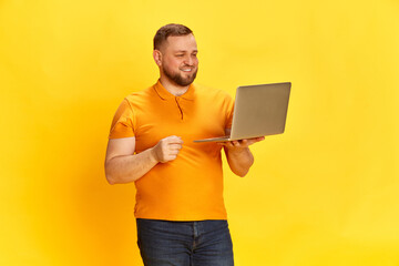 Smiling salesman holding laptop and working with pleasure over yellow background