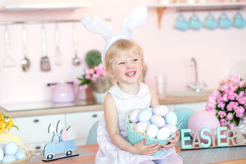 Fototapeta na wymiar Cute little girl with bunny ears holding Easter egg in the kitchen decorated for Easter.