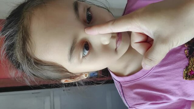 A little girl is practicing to do crossed eyes at home with an adult hand. Vertical video