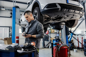 A car workshop worker is cleaning engine parts while standing at garage with car on a lift in a background.