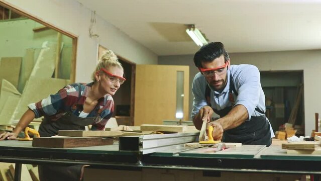 Two male and female carpenters work finishing wood furniture on table with electric cutting machine and female worker looking at attention.