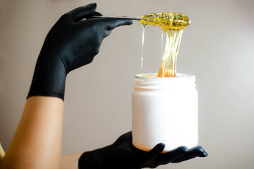 A female beautician holds a jar of shugaring sugar waxing paste, a light-coloured background.