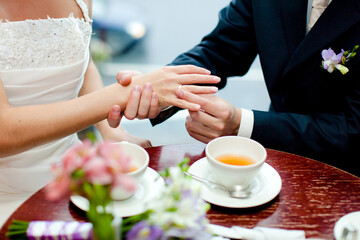 The groom puts the ring on the bride's finger at a table in a café. Only their hands are visible. There are flowers and cups of tea on the table.