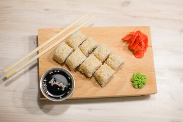 Traditional Japanese food. Freshly made sushi rolls of fish, rice, cheese, cucumber, sesame seeds served on a wooden board. Next to it: soy sauce, ginger, wasabi, chopsticks. 
