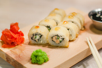 Traditional Japanese food. Freshly made sushi rolls of fish, rice, cheese, cucumber, sesame seeds served on a wooden board. Next to it: soy sauce, ginger, wasabi, chopsticks. 
