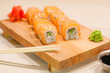 Traditional Japanese food. Freshly made sushi rolls of fish, rice, cheese, cucumber and sesame seeds served on a wooden board. Next to soy sauce, ginger, wasabi, chopsticks.