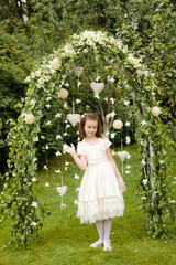 A girl in a smart white dress and gloves stands at a wedding flower arch in a park in summer. Full-length.