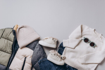 Fashion children's clothing, shoes - colorblock puffer jacket, knitted hat, scarf, boots, jeans, sunglasses on grey background. Spring, autumn collection. Top view, flat lay. Beige clothes outfit.
