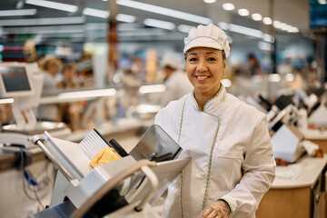 Happy supermarket worker using slicer at delicatessen section and looking at camera.