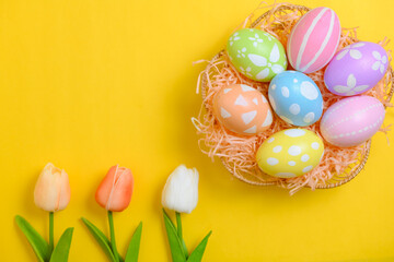Happy Easter holiday greeting card design concept. Colorful Easter Eggs and spring flowers on yellow background. Flat lay, top view, copy space.