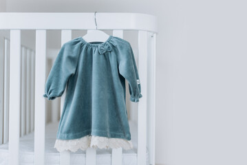 Blue dress for baby girl on white hanger near white bed in nursery. Set of baby clothes and accessories for birthday party. Fashion childs outfit. Copy space