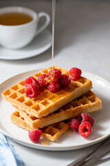 Pouring honey over sweet waffles with berries in plate served with cup of tea.
