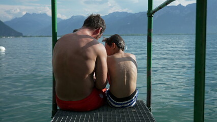 Father and son hanging out together by lake dock outside in scenice view of mountains in background. Summer family facations