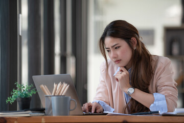 Sad Asian woman looking annoyed and stressed, sitting at the desk, using a laptop, thinking, feeling tired and bored with depression problems