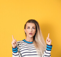 Young woman looking up and pointing to shows your space for product, promoter girl, isolated on yellow background