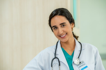Woman female doctor portrait in white coat gown with stethoscope sit and smile to front. Medical student attractive friendly wear white coat smiling and work on table in patient room