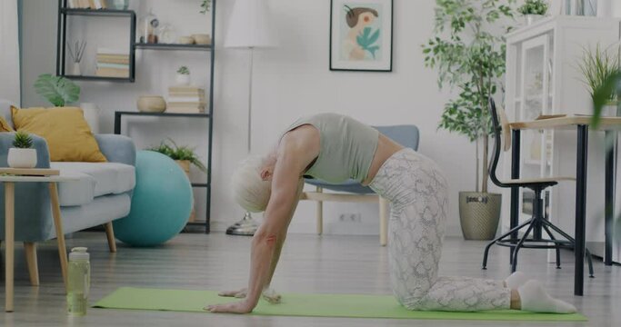 Side view of senior lady in sportswear stretching back caring for body health in living room at home. Active lifestyle and well-being concept.
