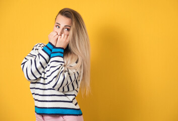 Worried young woman biting her fingernail, she know the secret, isolated on yellow background
