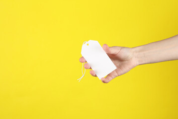 Female hand holds a white blank price tag on a yellow background. Sale concept