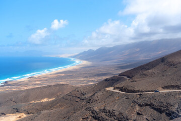 Fototapeta na wymiar Vista panorámica de la playa de Cofete con agua turquesa, arena blanca y algunas olas, rodeada de un paisaje volcánico y desértico durante un día soleado con cielo azul en Fuerteventura, Isla Canaria