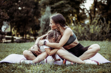 Athletic mom hugs her little daughter while exercising on the mat in the park. Healthy family concept, motherhood